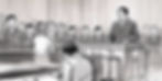 ชายยืนพูดในห้องพิจารณาคดี คนอื่นๆ ฟังอย่างตั้งใจ บรรยากาศเคร่งขรึม พื้นหลังเป็นม่านบางๆ ขาวดำ