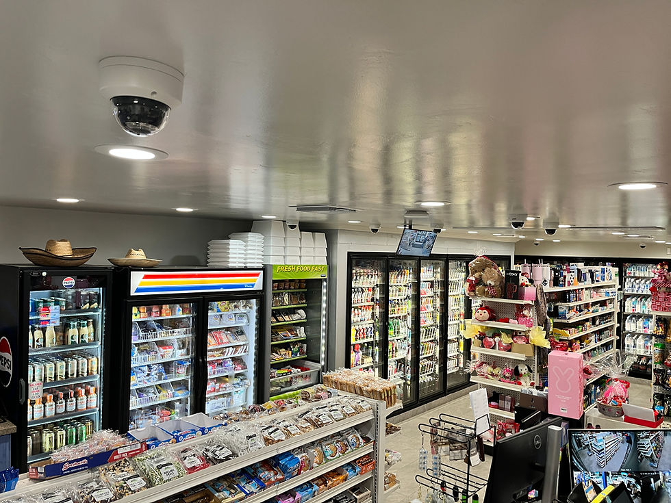 Convenience store aisle with snack racks, drink coolers, gift displays, and ceiling security cameras under bright lights
