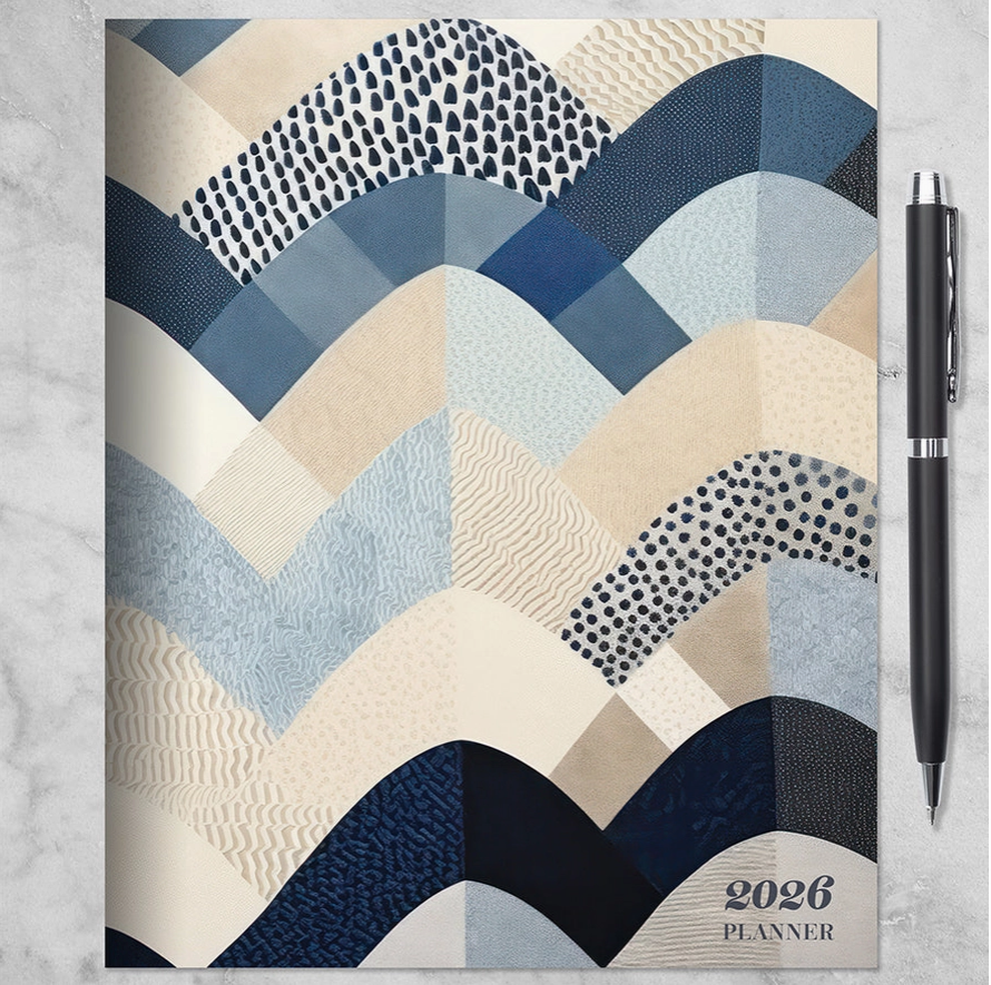 2026 Quilted Curves monthly planner with blue, cream, and navy patchwork style cover and black pen beside it.”