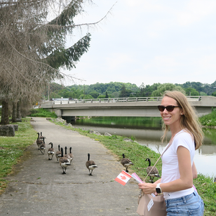 Viktoria Gilanyi of ORIA Interiors holding a Canadian flag while walking along a riverside path on Canada Day, reflecting on home and belonging.