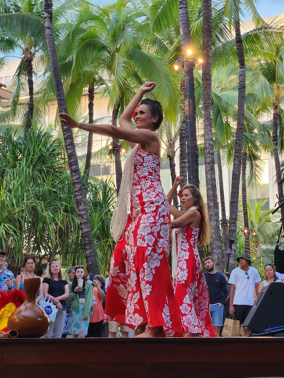 Two women in red floral dresses dance the hula on an outdoor stage among palm trees, with a small crowd watching in a tropical setting.