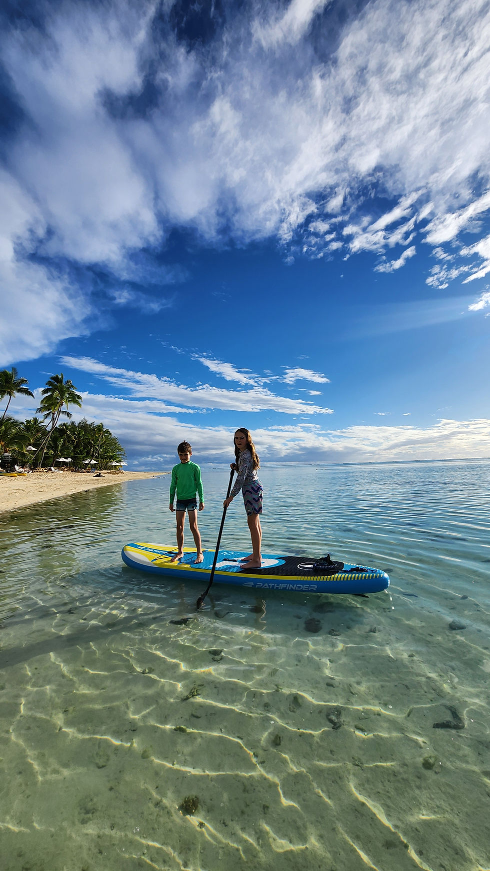 Two kids on a paddleboard in clear water, one paddling. Sunny beach with palm trees in the background, blue sky with clouds above in Aitutaki, Cook Islands.