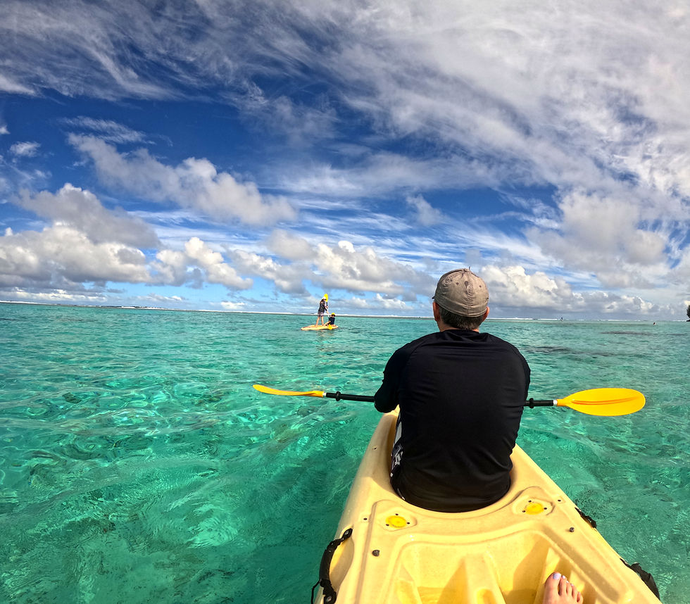 Man kayaking in clear turquoise ocean, with children paddleboarding in the distance under a bright blue, cloudy sky in Aitutaki. Cook Islands.