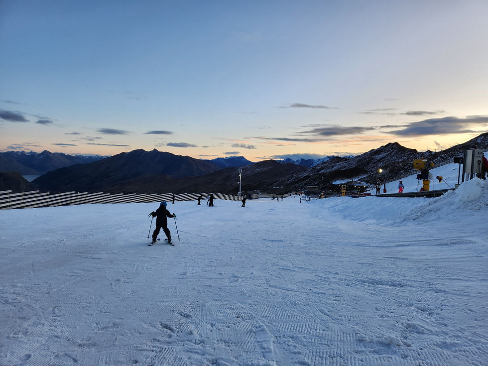A skier descends a snowy slope at sunset, with mountains in the background. Other skiers and ski equipment are visible under a colorful sky.