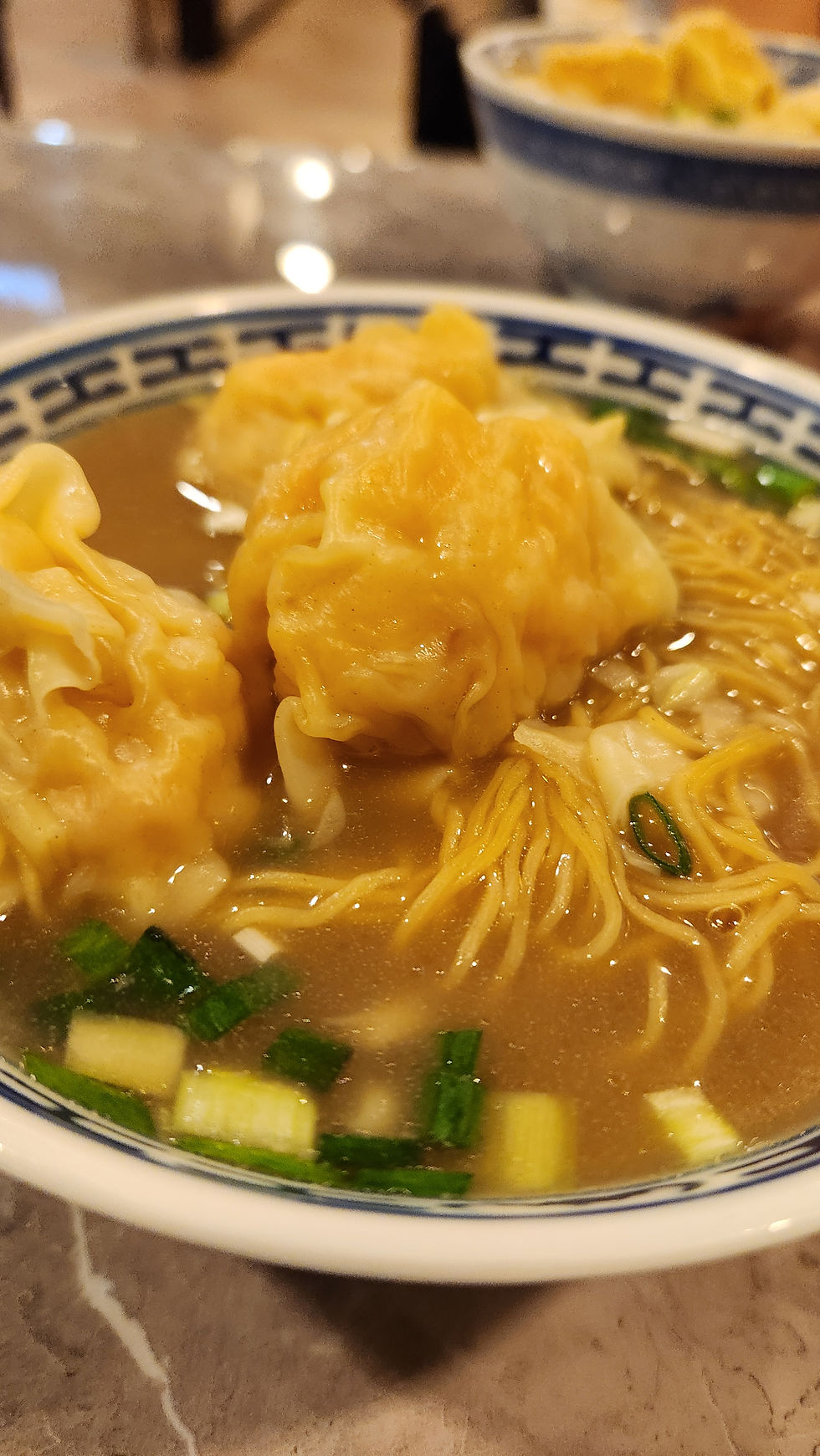 Bowl of wonton noodle soup with yellow dumplings, scallions, and broth on a restaurant table, warm and appetizing