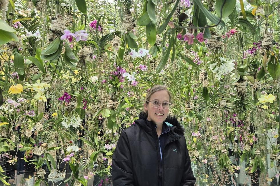 Woman in a black jacket smiles in a lush greenhouse with hanging orchids in pink, purple, and yellow hues, creating a vibrant backdrop at teamLab Planets Toyosu, Tokyo, Japan
