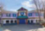 Purple building labeled Village Shop with green roof, blue windows, trees nearby. Signs read Toys, Gifts, Souvenirs. Bright day.