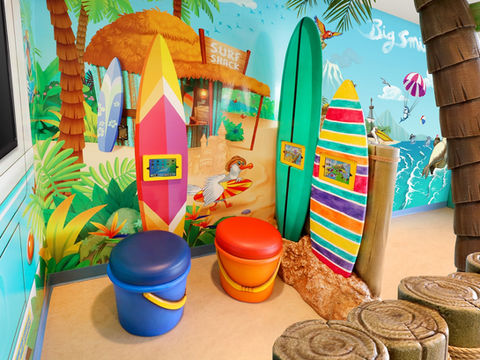 Surf-Themed Interactive Play Area: A beach and surf-themed play area featuring colorful surfboards and tablet stations, set against a tropical mural backdrop for a fun, interactive experience for kids.