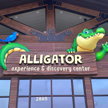 Alligator Experience & Discovery Center: A playful alligator peeking over a wooden sign with bright colors, inviting guests to explore the center.
