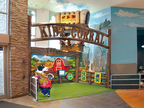 Western Cowboy Corral Play Area: A western-themed indoor play area called "Kiddie Corral" with a barnyard background and a cartoon cow, offering interactive games and a ranch-style play environment.