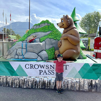 Crowsnest Pass Parade Float: A whimsical float with a large bear sitting beside a stream and a fish, with a child posing in front, under the banner "Crowsnest Pass."