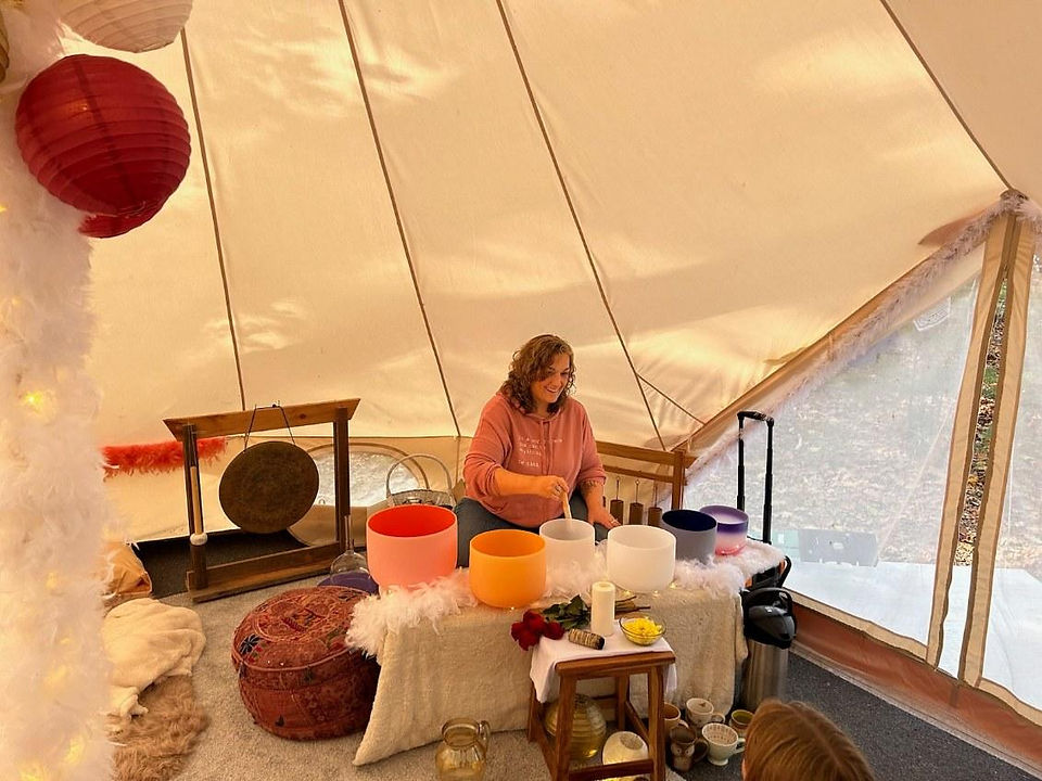 inside a white dome tent. table with glass singing bowl in front of Kristen who is playing them and smiling