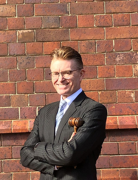 Phil Brogan in a tuxedo, holding a gavel, stands confidently, ready to preside over a formal event.