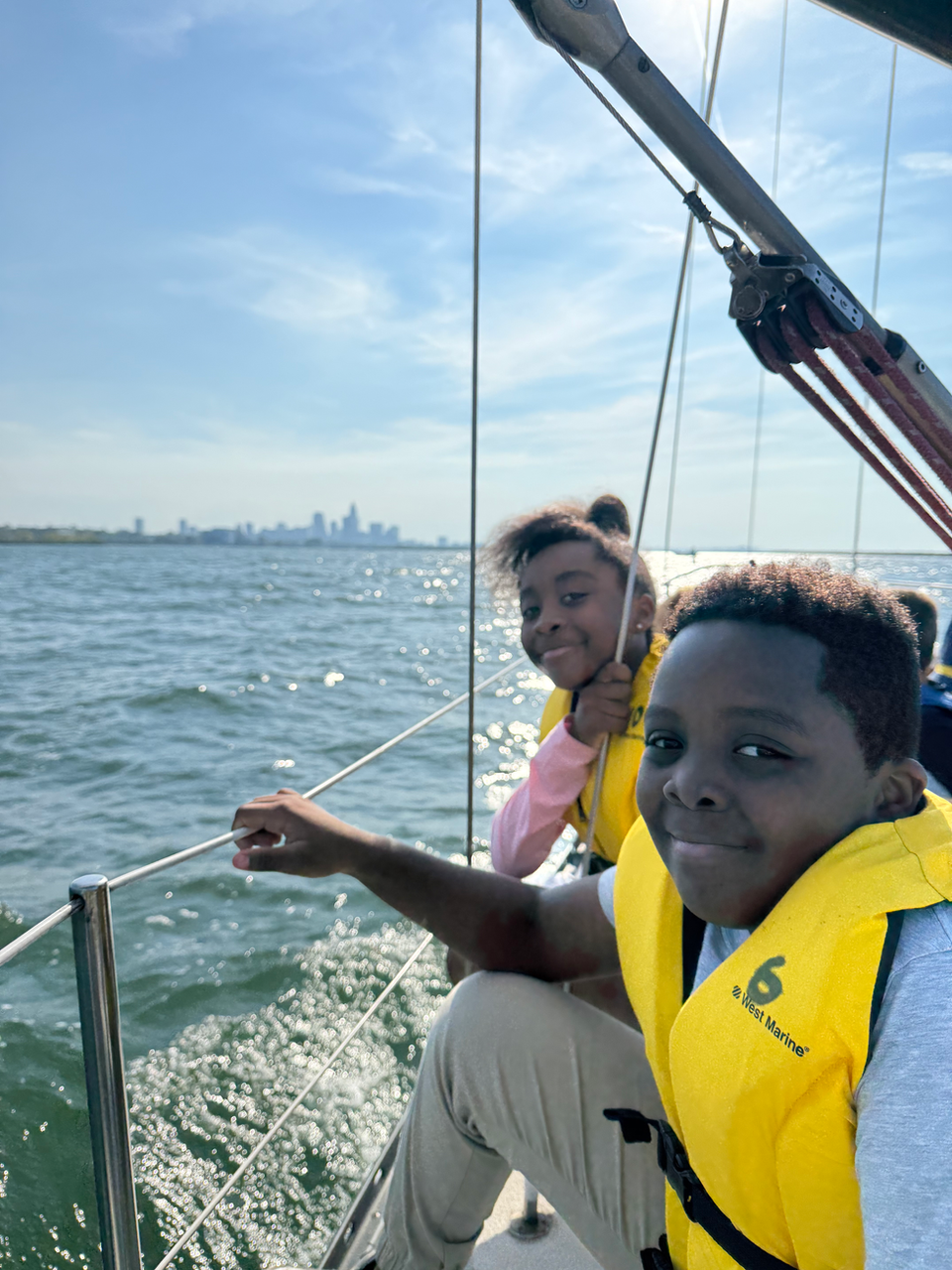 Smiling children in life jackets on a sailboat with city skyline.