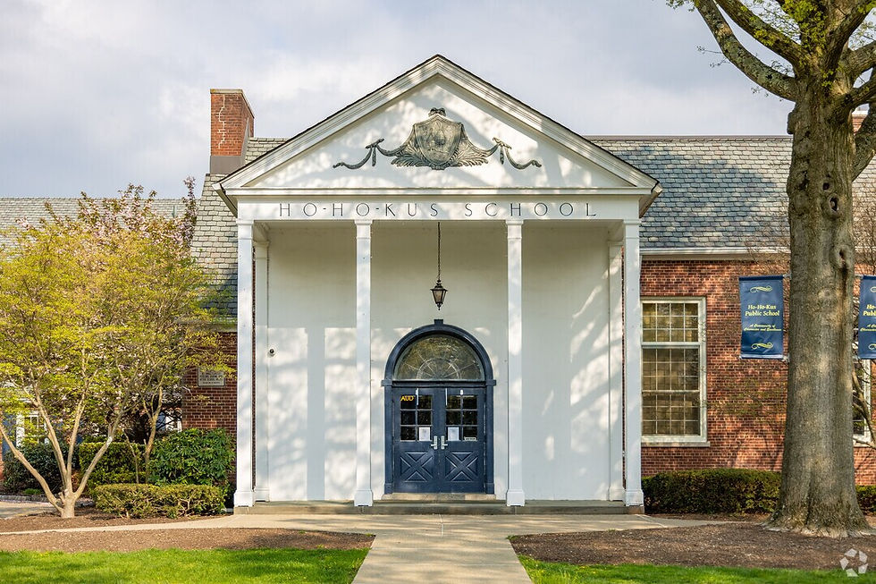 White facade of Ho-Ho-Kus School with columns and dark blue arched door, surrounded by brick walls, trees, and a path. Bright, inviting atmosphere.