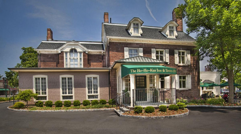 Historic two-story brick inn with white trim, green awning reads "The Ho-Ho-Kus Inn & Tavern." Surrounded by landscaping under a clear blue sky.
