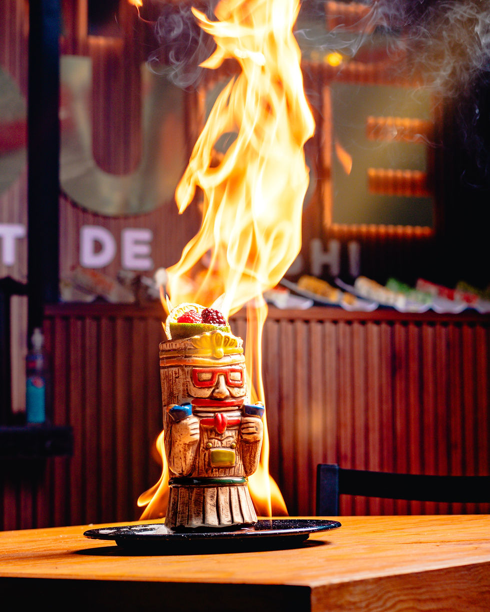Flaming tiki cocktail in a carved tiki mug on a wooden table, with orange flames and smoke in a dim bar.