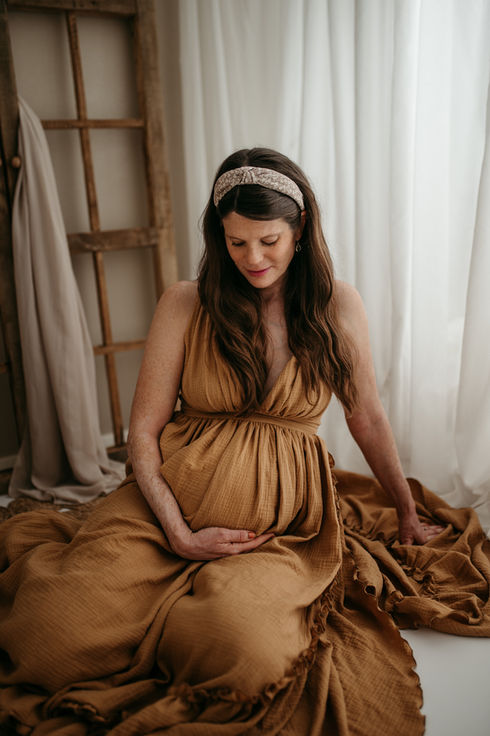 Junge, werdende Mama sitzt am Boden, vor einem Vorhang, in einem wunderschönen Kamelfarbenen Schwangerschaftskleid. Sie schaut zum Bauch und sieht glücklich aus. 