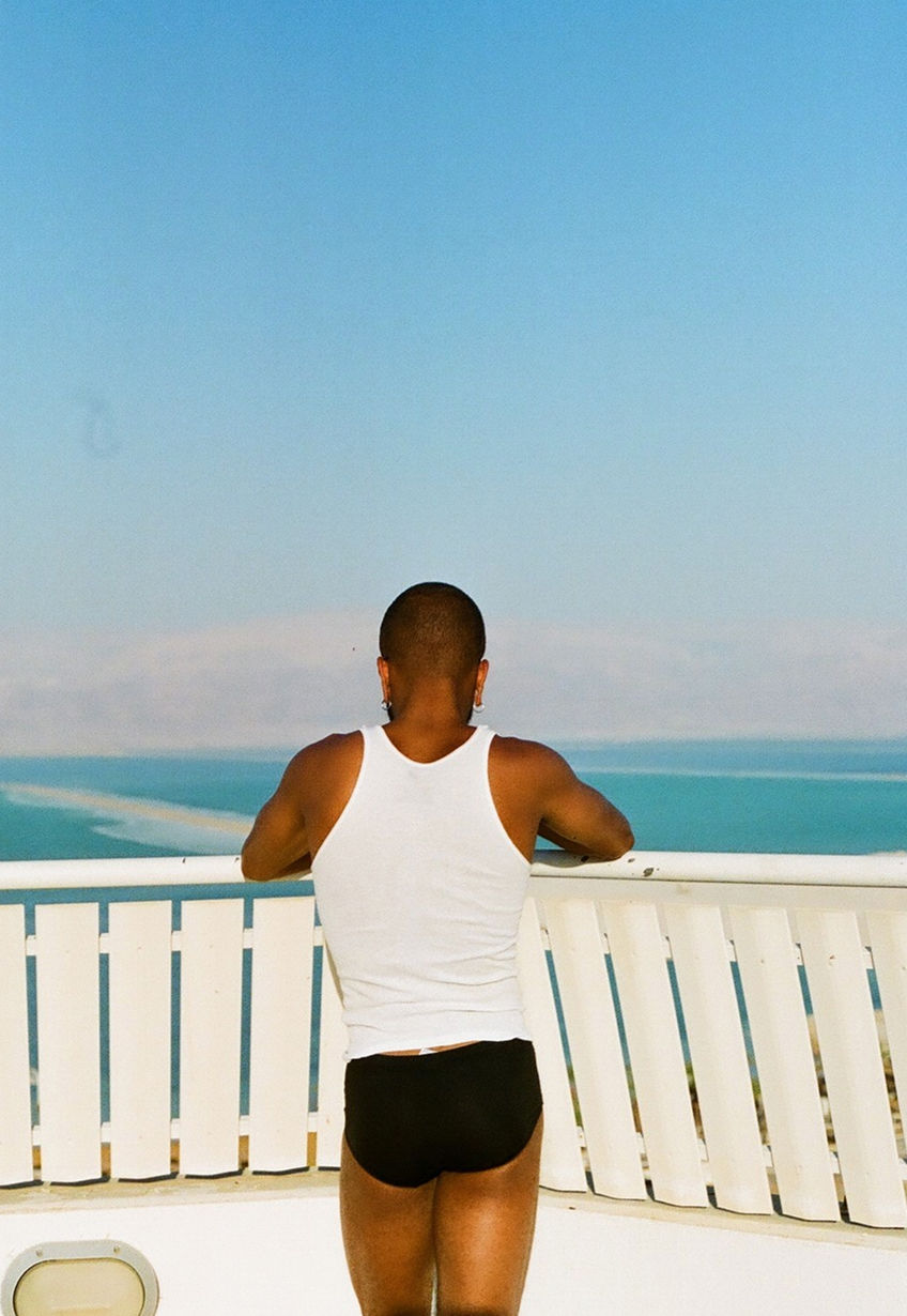 A man is seen from the back wearing a white tank top and dark swimwear. He is posed at a white railing overlooking a sea under a bright blue sky.