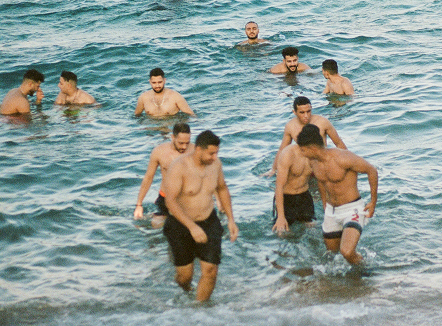 The image shows a group of men in the ocean. Some are deeper in the water, up to their torso, while others are closer to the shore, wading out. The water is a greenish-blue color. 