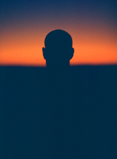 A silhouette of a bald person against an orange sunset. The person's head and shoulders are visible above a dark blue horizon line. 