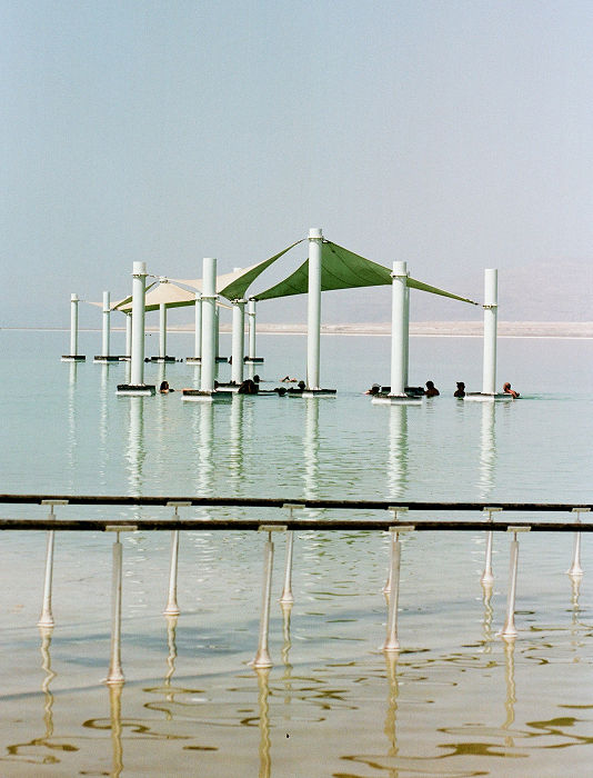 A vertical shot depicts a serene scene at the Dead Sea. In the distance, a line of white posts supports canvas sunshades over people bobbing in the water.