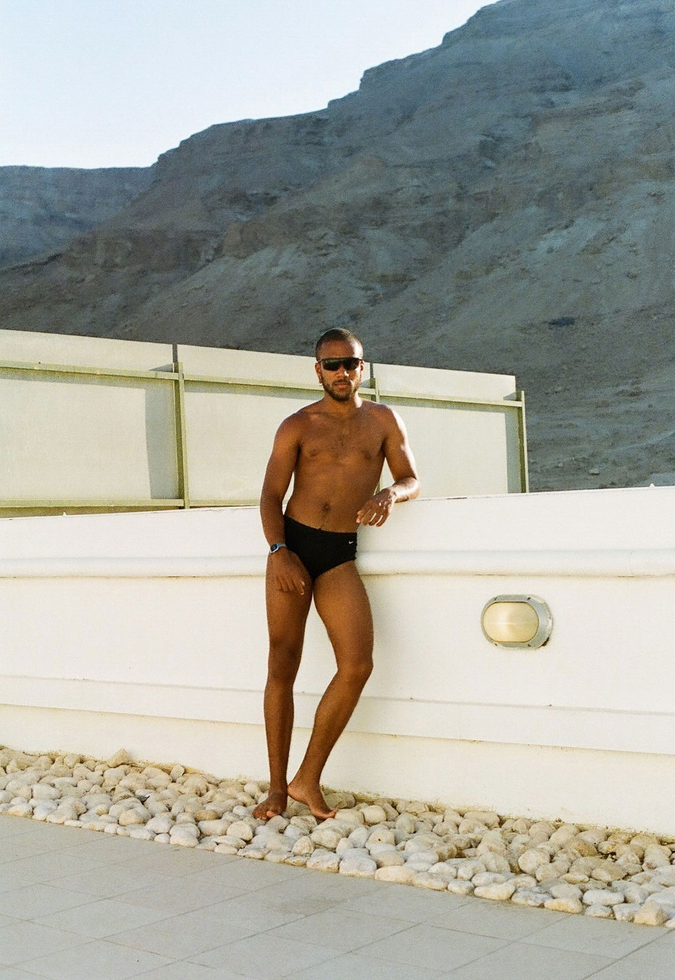 A medium shot presents a shirtless man in a suit against a bright, sunny landscape. The man, with dark hair/facial hair and sunglasses, leans casually against a white wall, wearing a watch and dark swimwear.