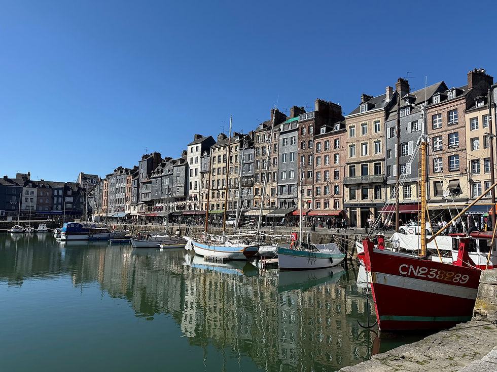 Boats docked in a tranquil harbour with colourful historic buildings reflecting in the water under a clear blue sky.
