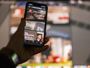 Eine Hand die ein Handy mit der geöffneten neuen Webseite zeigt. Im Hintergrund die Webseite auf einem PC.