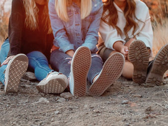 Three people are sitting on the ground with their legs stretched out in front of them. They are wearing casual clothing, jeans and sneakers and are surrounded by dry grass and dirt. The setting appears to be outdoors, likely at sunset.