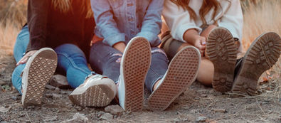 Three people are sitting on the ground with their legs stretched out in front of them. They are wearing casual clothing, jeans and sneakers and are surrounded by dry grass and dirt. The setting appears to be outdoors, likely at sunset.