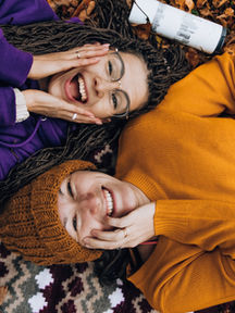 Pexels photo - Two people smile as they lie on the ground covered with autumn leaves. One wears an orange sweater and matching hat, while the other wears a purple sweatshirt. They both touch each other's cheeks in a cheerful and relaxed manner, with a white thermos beside them.