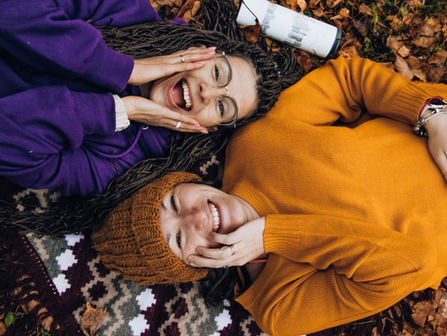 Pexels photo - Two people smile as they lie on the ground covered with autumn leaves. One wears an orange sweater and matching hat, while the other wears a purple sweatshirt. They both touch each other's cheeks in a cheerful and relaxed manner, with a white thermos beside them.