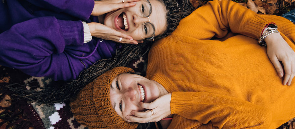 Pexels photo - Two people smile as they lie on the ground covered with autumn leaves. One wears an orange sweater and matching hat, while the other wears a purple sweatshirt. They both touch each other's cheeks in a cheerful and relaxed manner, with a white thermos beside them.