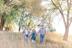 Three young children holding hands and walking outdoors in a field with trees.