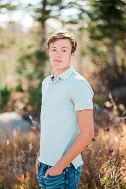 A young man in a light blue polo shirt standing outdoors against a backdrop of rocks and greenery.
