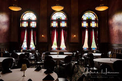 Blurred interior view of a wedding reception in a grand hall with arched windows and chandeliers.