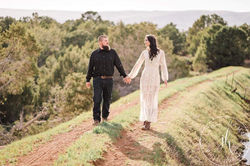 A couple holding hands and walking away on a dirt path in a hilly landscape.