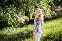 A young woman with long blonde hair wearing a patterned top and jeans standing in a grassy area.