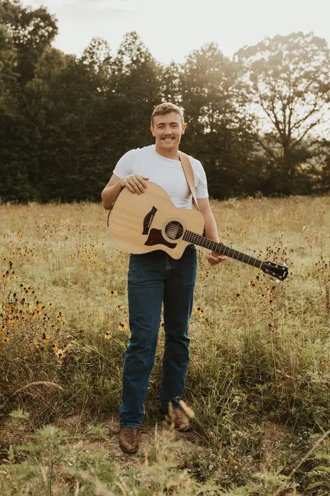 Braden Wall standing in an open field in a white t-shirt holding his guitar