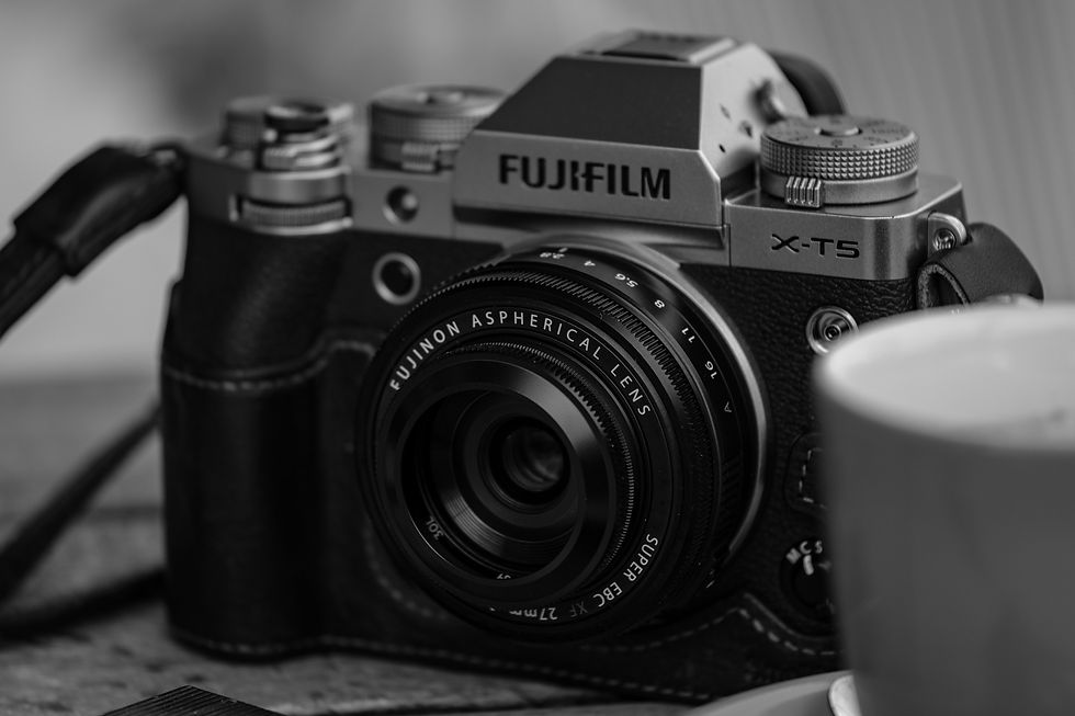 Black-and-white close-up of a Fujifilm X-T5 camera with Fujinon lens on a table, with straps and a cup nearby.