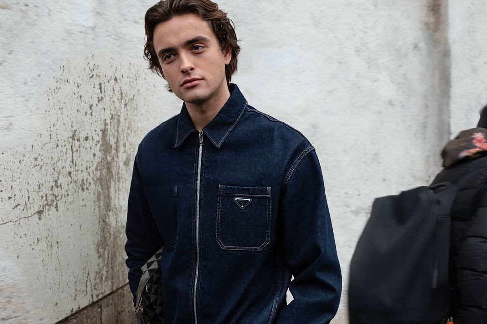 Young man in a dark denim jacket stands against a stained white wall, looking serious and thoughtful.