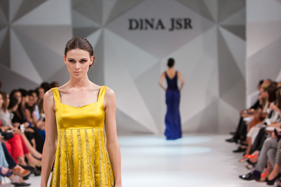 Fashion runway model in yellow dress at DINA JSR show, with audience seated on both sides and a blue-gowned model in back