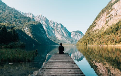person seated at end of a pier overlooking water and mountains