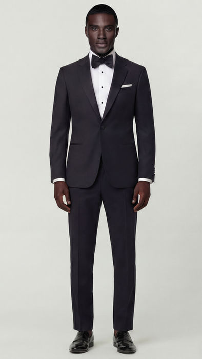 A well-dressed man in a classic black notch tuxedo with a white dress shirt, black tie, and white pocket square standing confidently beside a woman in an elegant white gown, both smiling warmly against a light grey background.