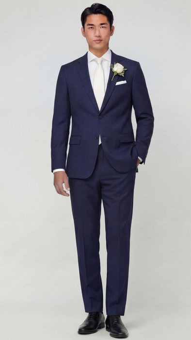 A man dressed in a notch lapel navy blue suit with a white dress shirt, navy striped tie, and white pocket square, standing closely beside a woman in a strapless white gown, both elegantly styled and posing confidently against a light grey background.