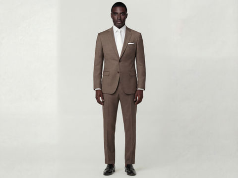 A man wearing a notch lapel mid-brown suit with a subtle birds-eye pattern, white dress shirt, white tie, and white pocket square, posing with a confident expression against a light grey background.