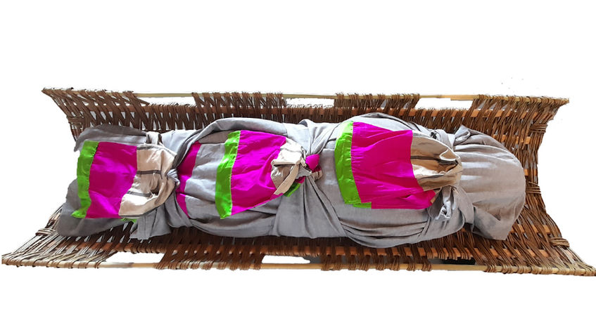Grey burial shroud wrapped with three bright pink and green fabric ties inside a willow stretchers for natural burial.