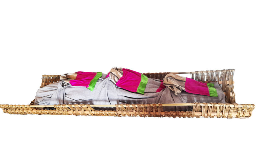 Grey burial shroud wrapped with three bright pink and green fabric ties inside a willow stretchers for natural burial.