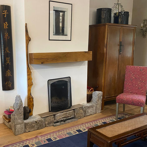 Sitting room hearth in a restored 1670 granary, County Mayo. A cast iron fireplace framed by rough stone, an oak beam mantel above, a tall antique wooden cabinet, a pink floral chair, and a Chinese calligraphy panel on the wall.